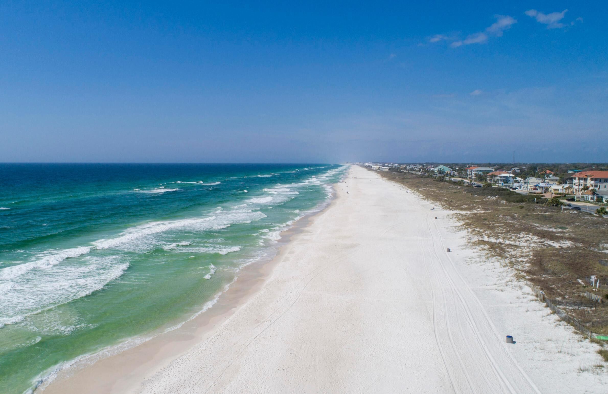 Une vaste plage de sable fin, bordée par l’océan et ses vagues, sous un ciel dégagé, avec des bâtiments qui s’alignent le long du rivage.