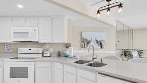 A modern kitchen with white cabinetry, a built-in microwave, stove, and dishwasher. There is a large island with a sink and a pendant light fixture above. - Aquavista 404e (Panama City Beach)