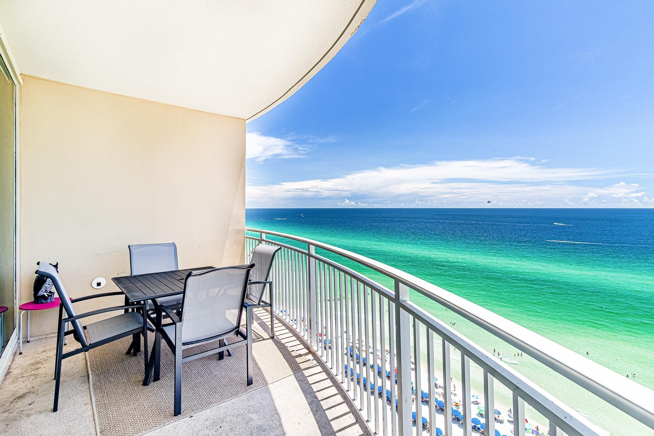 A balcony with a table and chairs overlooking a beach with clear turquoise water and a few boats.