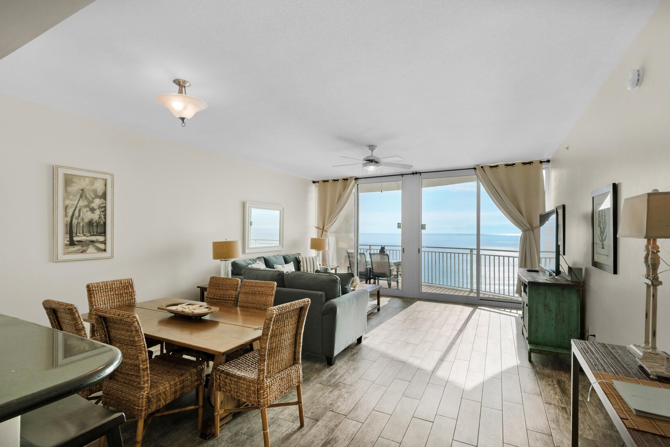 A spacious living area with a dining table, wicker chairs, a sofa, and a balcony with a view of the sea.