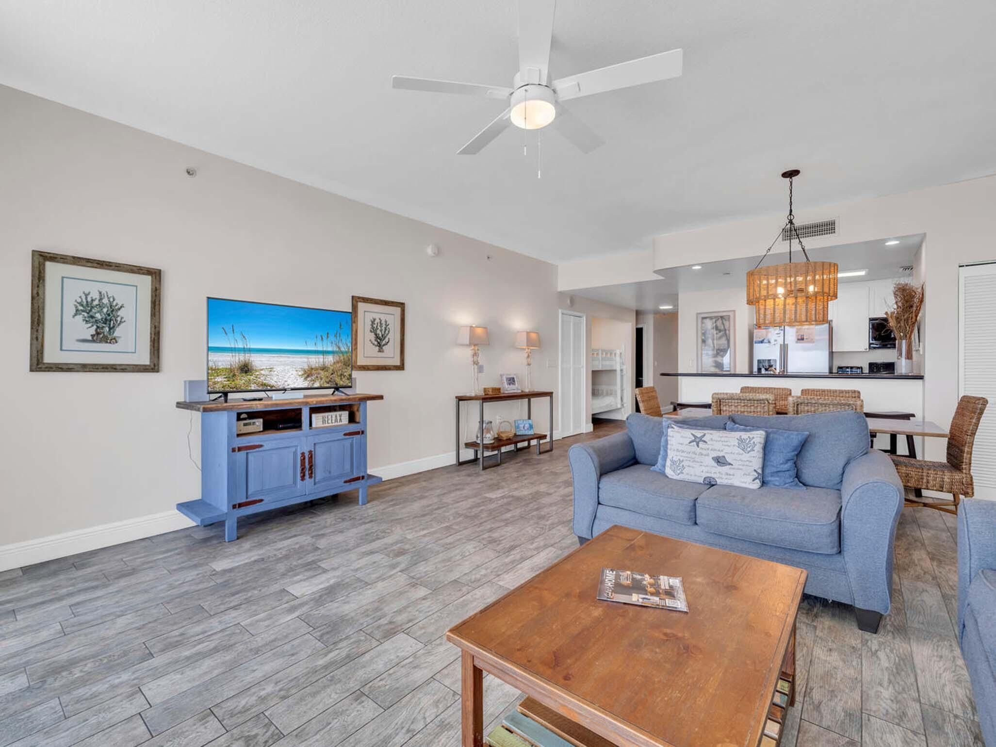 A spacious living room with a blue sofa, a coffee table, and a TV on a wooden stand. There is a ceiling fan, a dining area with wicker chairs, and a kitchen in the background.
