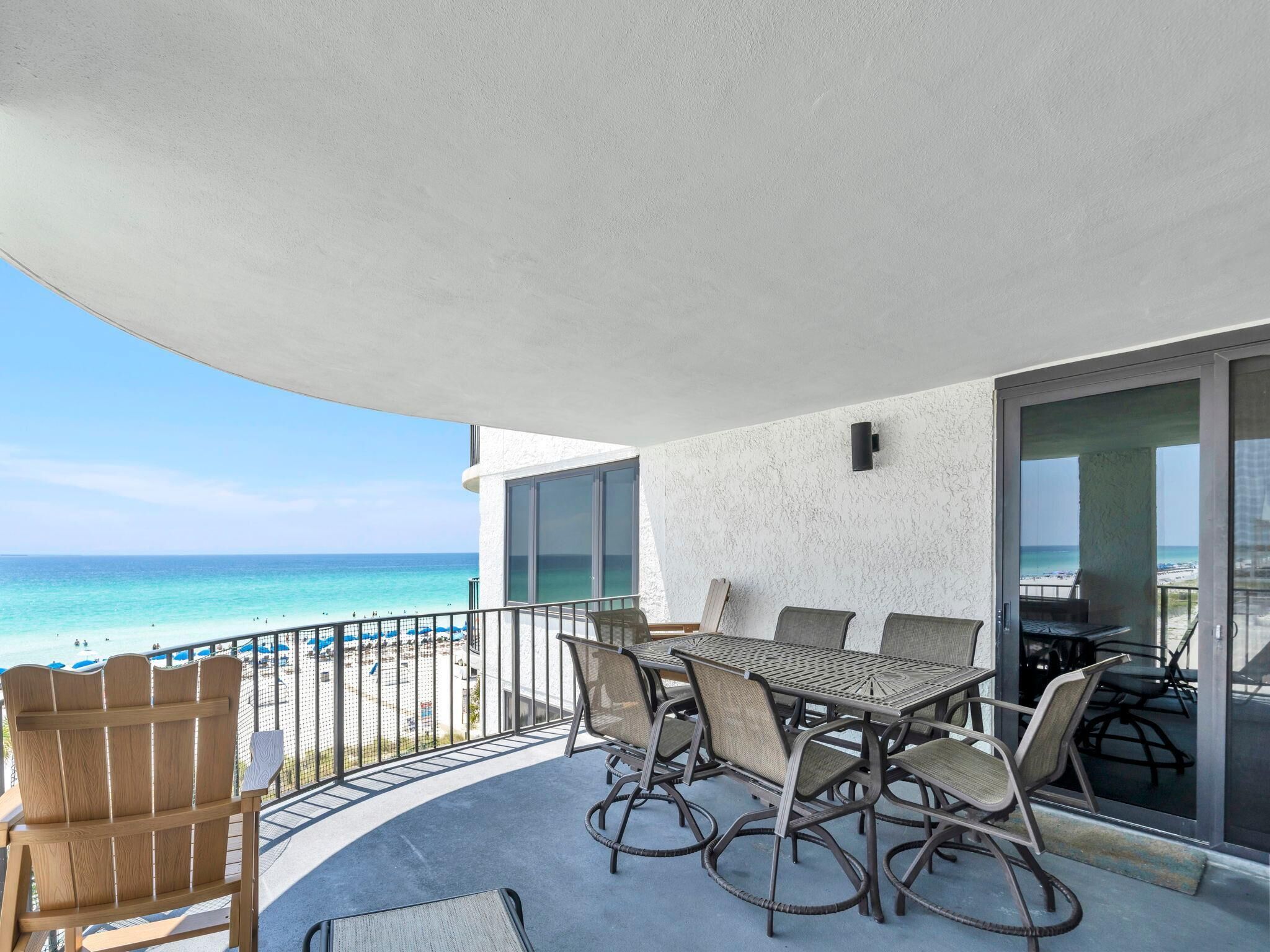 Een balkon met een tafel en stoelen, met uitzicht op een strand met mensen en parasols.