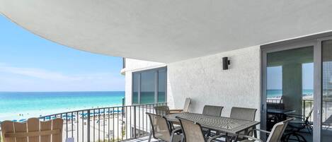 Een balkon met een tafel en stoelen, met uitzicht op een strand met mensen en parasols.