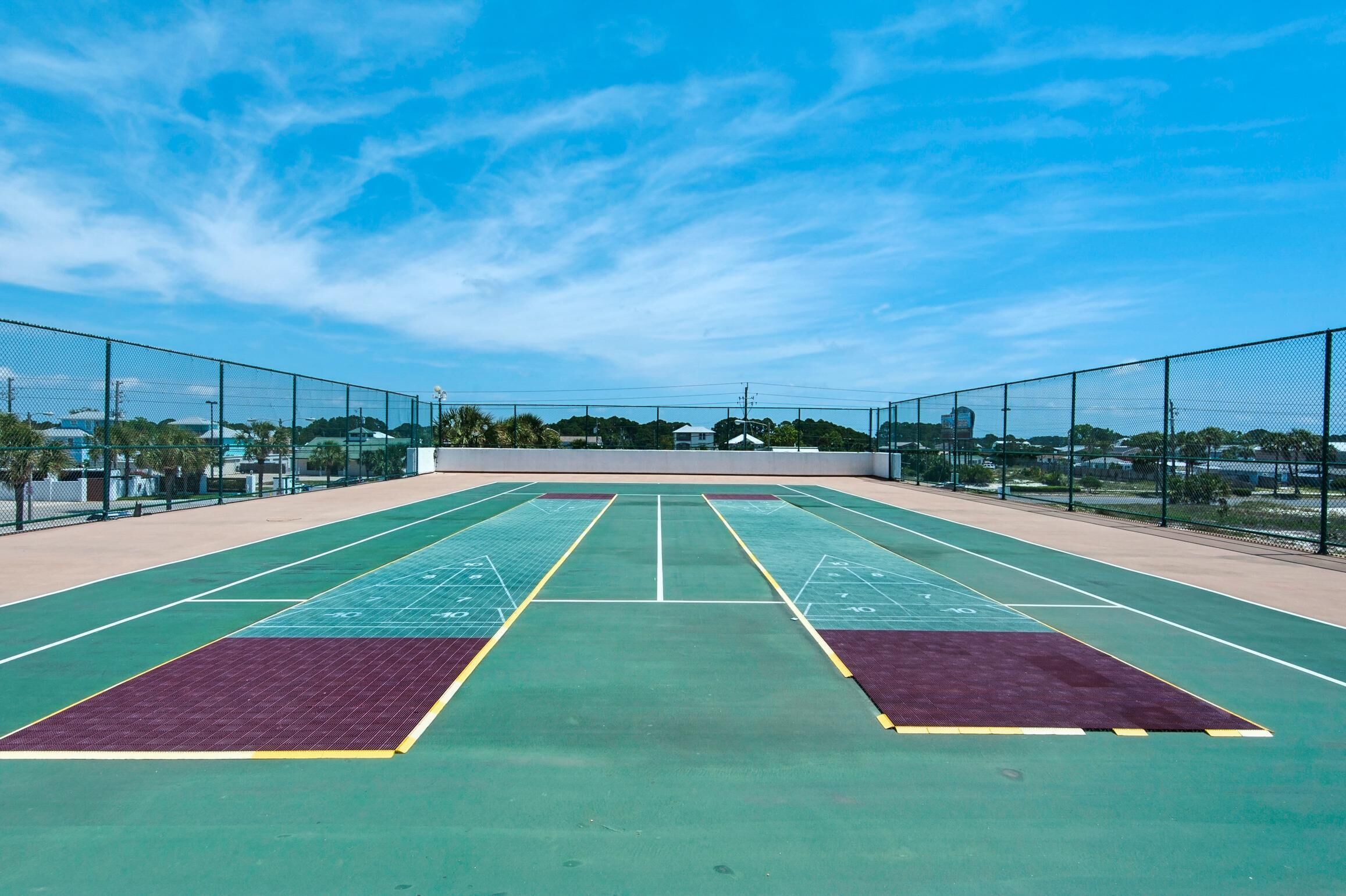 An outdoor tennis court with a net, surrounded by a fence, and a clear sky above.