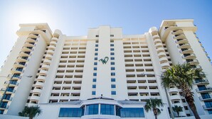 Un hôtel à plusieurs étages doté d’une entrée centrale, de balcons à chaque étage et de palmiers à l’avant.