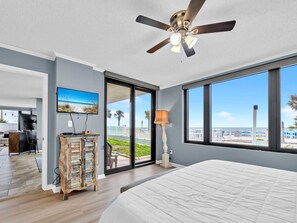 A bedroom with a large bed, a ceiling fan, a TV, and a view of the beach through a sliding glass door.