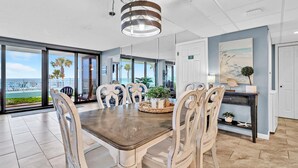 Un vaste espace dédié à la salle à manger, avec une grande table et des chaises, un luminaire suspendu et une vue sur l’océan grâce à de grandes fenêtres.