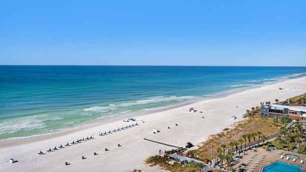 A beach resort with a long stretch of beach, a pool area, and a building with a blue roof. - The Summit 1432 (Panama City Beach)