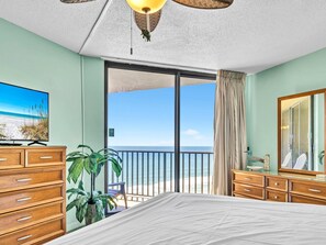 Een hotelkamer met een bed, een houten dressoir, een televisie waarop een strand te zien is, een balkon met uitzicht op de oceaan, en een grote spiegel.