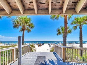Een houten terras dat uitkomt op een strand, met palmbomen en blauwe parasols.