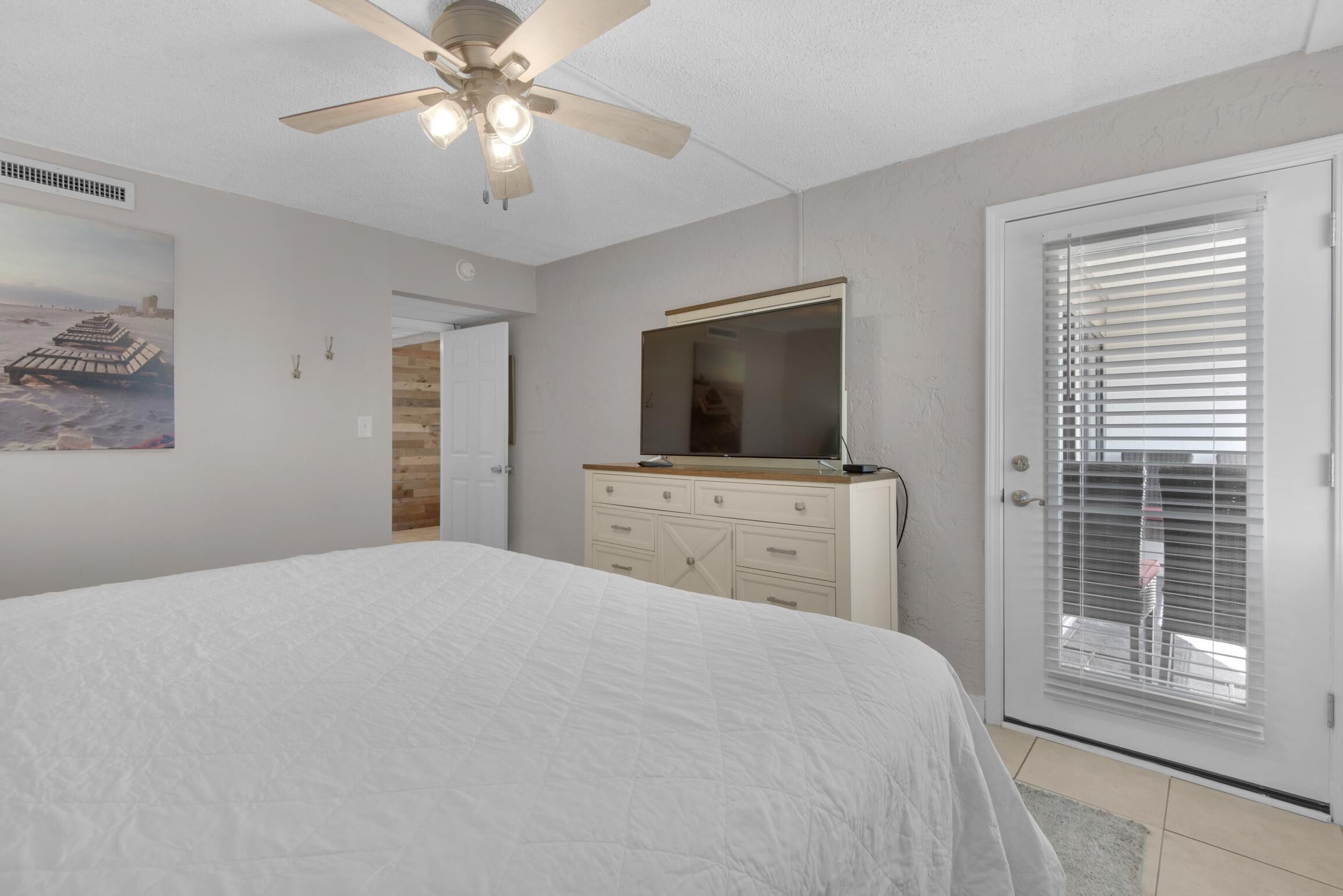 A bedroom with a bed, a dresser, a ceiling fan, a door with blinds, and a wall-mounted mirror.