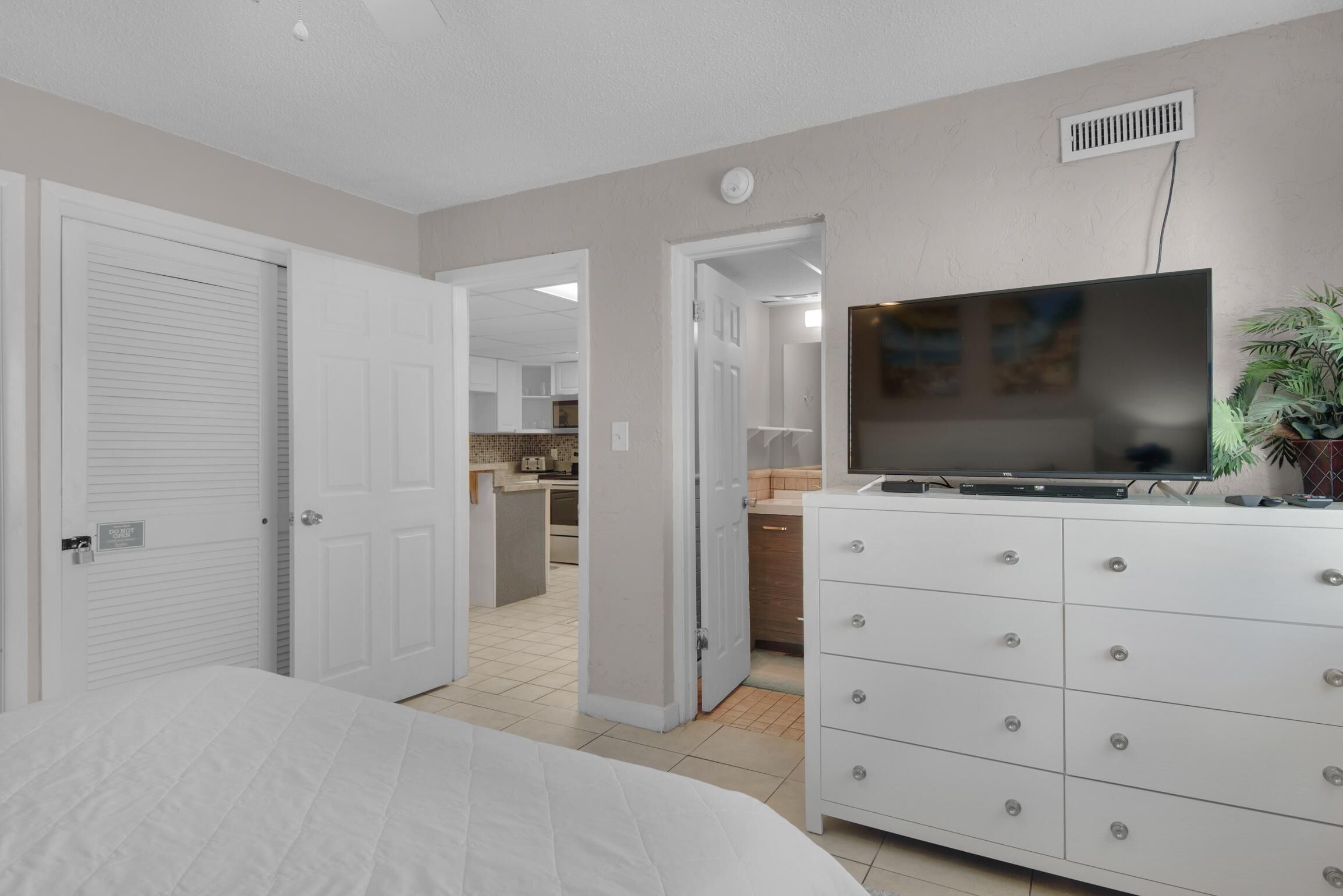 Un dormitorio con una cama, un televisor sobre un aparador y una cocina al fondo.