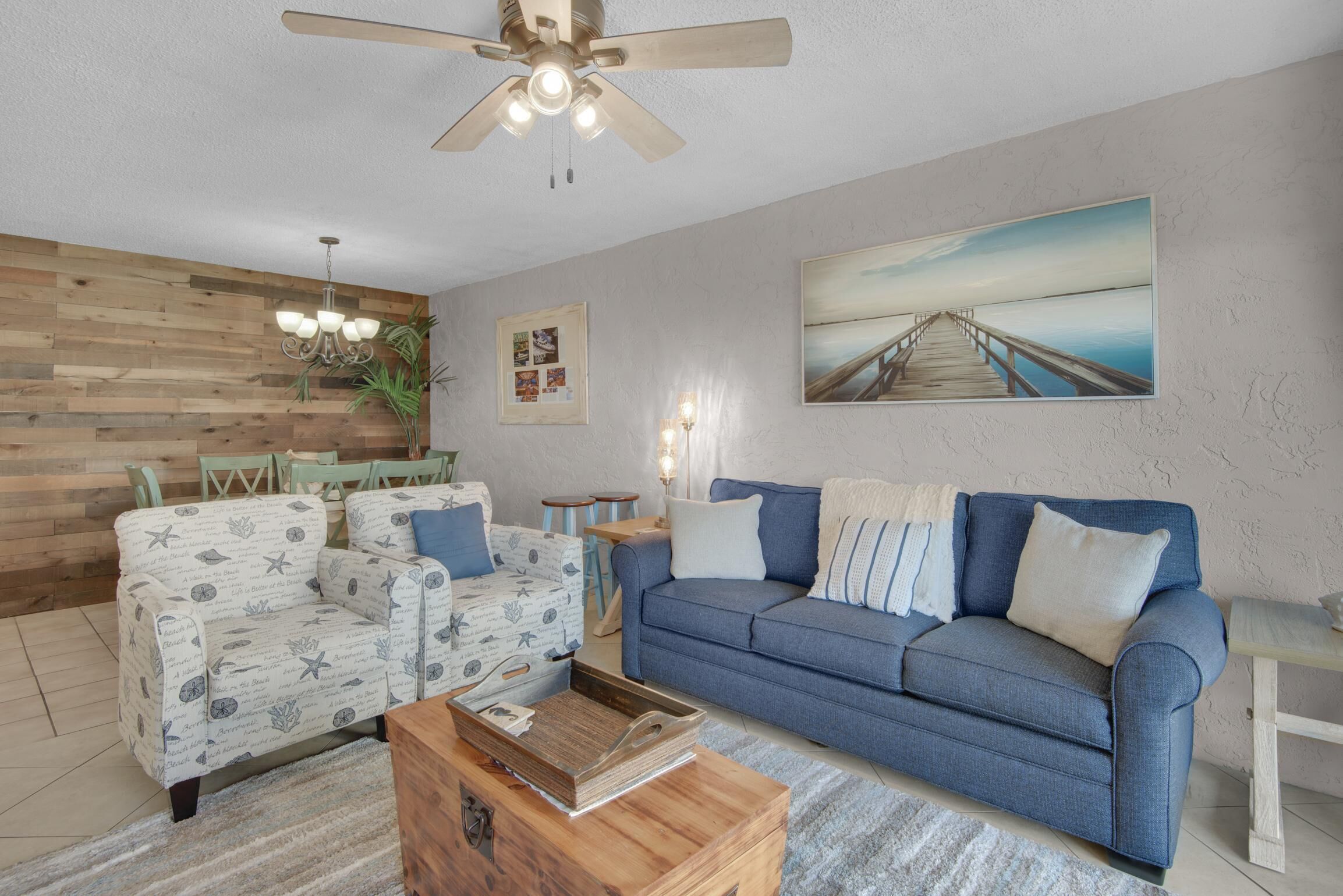 A living room with a wooden wall, a blue sofa, a patterned armchair, a coffee table, and a ceiling fan.