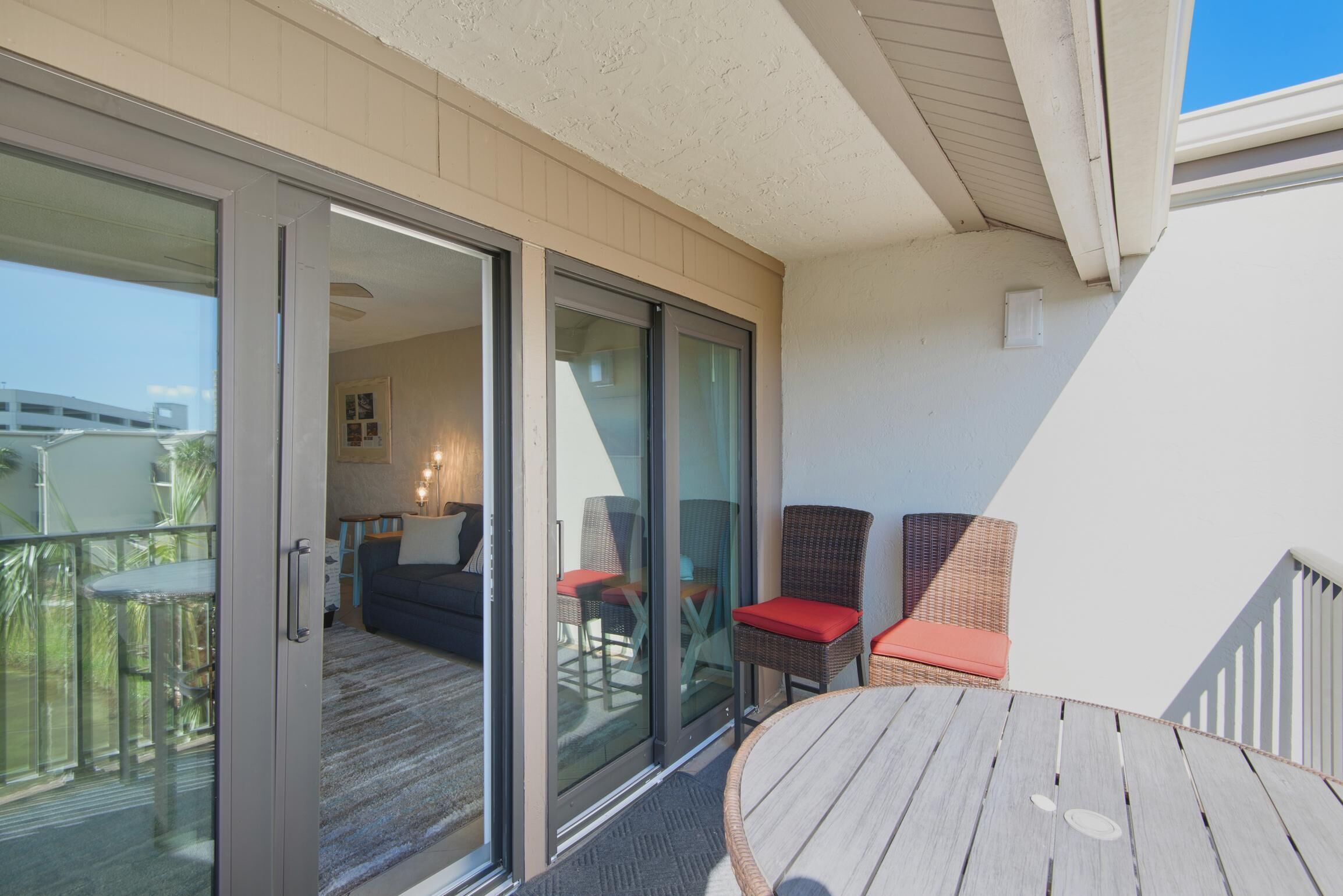 A balcony with a sliding glass door, wicker chairs with red cushions, and a wooden table.