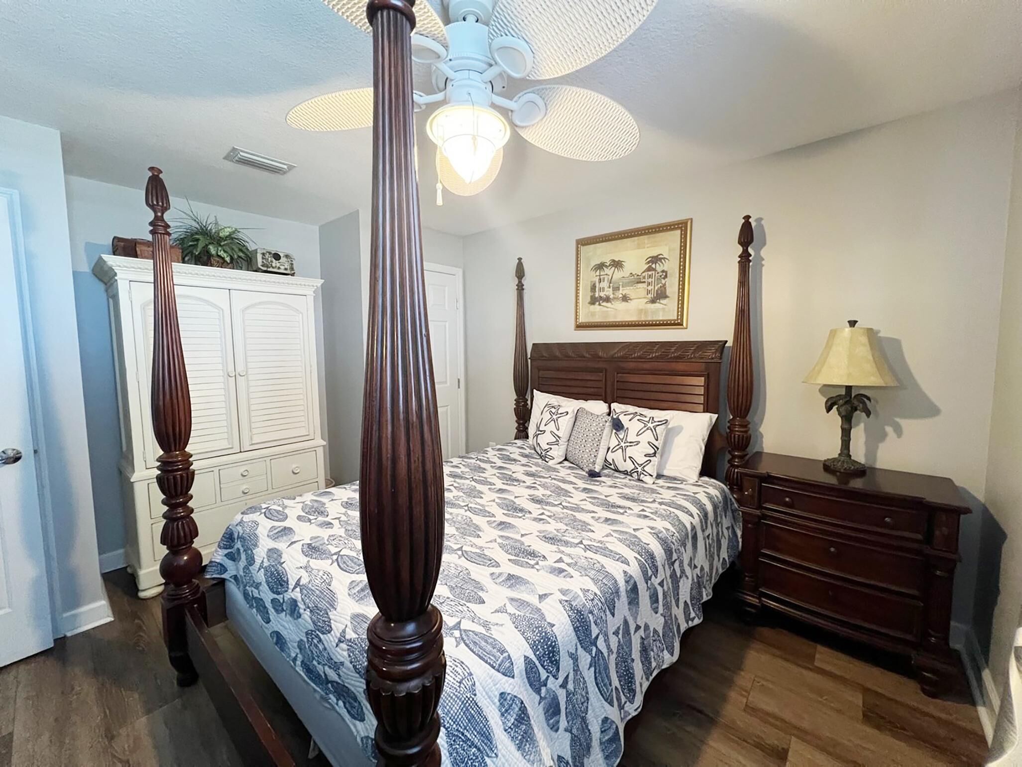 A four-poster bed with a patterned bedspread, a nightstand with a lamp, a ceiling fan, and a framed picture on the wall.