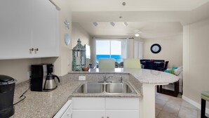 A kitchen with a coffee maker, double sink, and cabinets. There is a view of the ocean through a window.