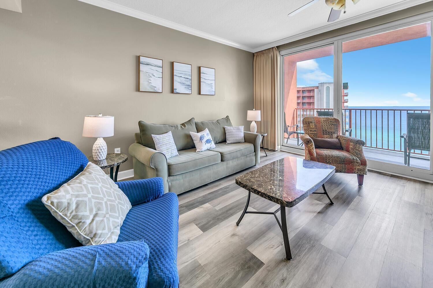 Uma sala de estar moderna com um sofá cinzento, uma poltrona azul e outra com padrão, uma mesa de centro de mármore e uma grande janela com vista para o mar.