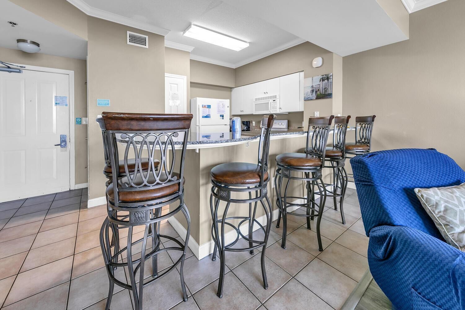 A kitchen with a white refrigerator, microwave, and stove. There are four bar stools with metal frames and wooden seats. A blue couch is in the foreground.