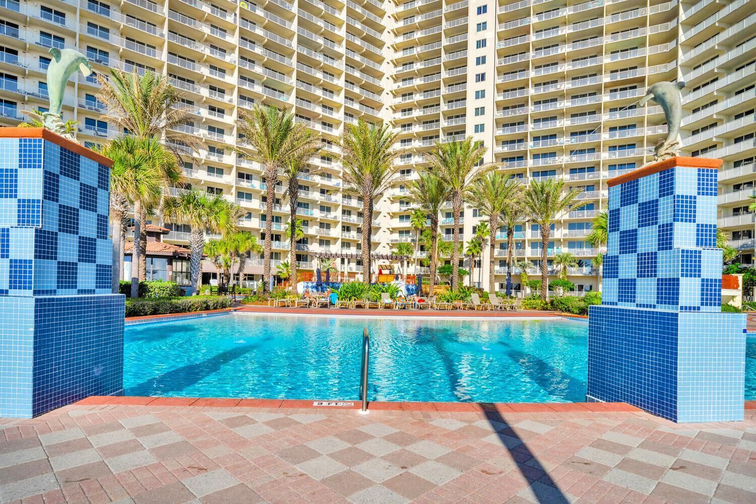 A hotel pool area with a large pool, palm trees, and a high-rise building in the background.