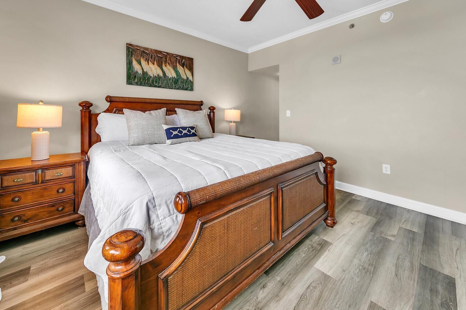 A bedroom with a wooden bed, two nightstands, a painting on the wall, and a ceiling fan.