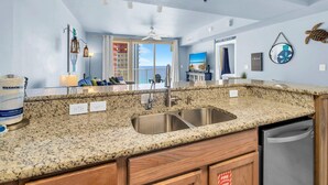 A kitchen with granite countertops, a stainless steel sink, wooden cabinets, and a view of the ocean through large windows. - Shores of Panama 1816 (Panama City Beach)