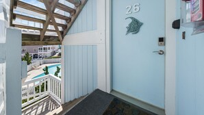 Eine hellblaue Tür mit der Nummer 26 und einem Fischemblem, ein Holzbalkon mit Blick auf einen Pool und weitere Gebäude sowie ein an der Wand befestigter Feuerlöscher.