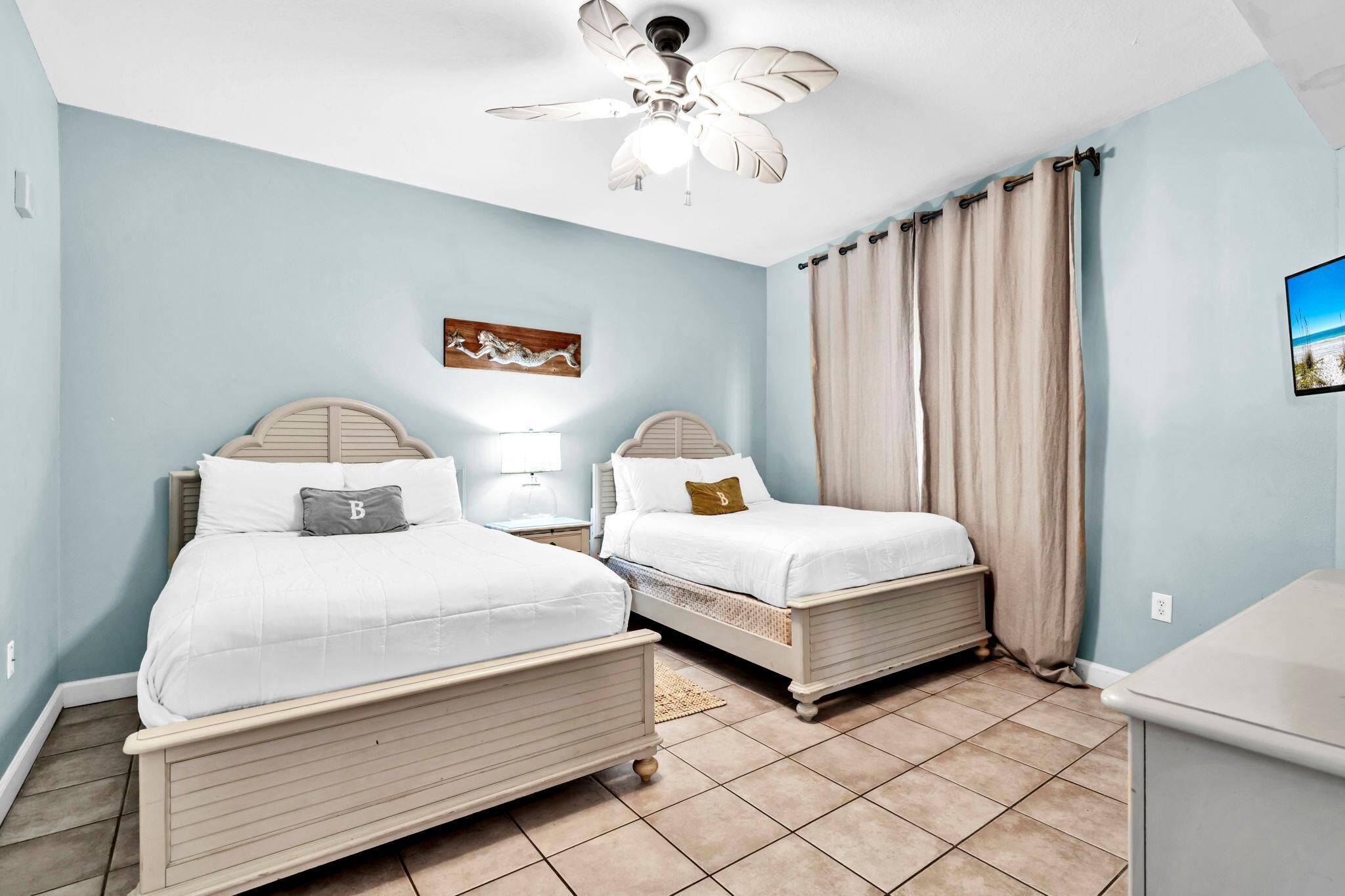 Un dormitorio con dos camas, un ventilador de techo, un cuadro y una ventana con cortinas.