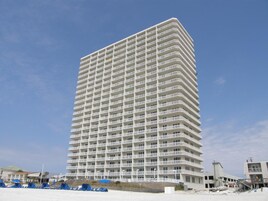Un hotel alto y rectangular con varios balcones, situado cerca de una playa con algunas estructuras azules al frente.