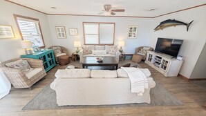 Een woonkamer met een grote beige bank, twee rieten fauteuils, een salontafel en een televisie. Er hangen ingelijste foto's aan de muren en er is een plafondventilator.