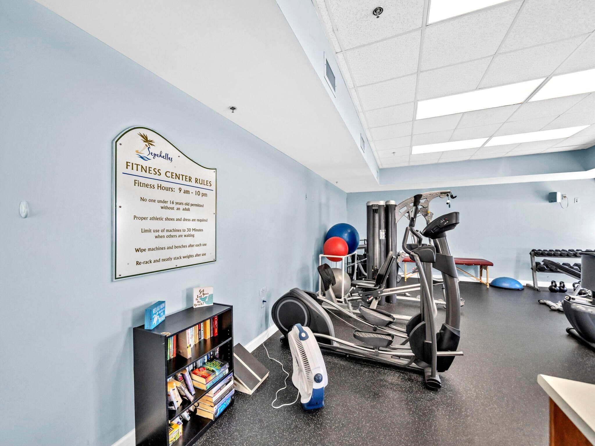 A fitness center with exercise equipment, a sign detailing rules, and a bookshelf with books.
