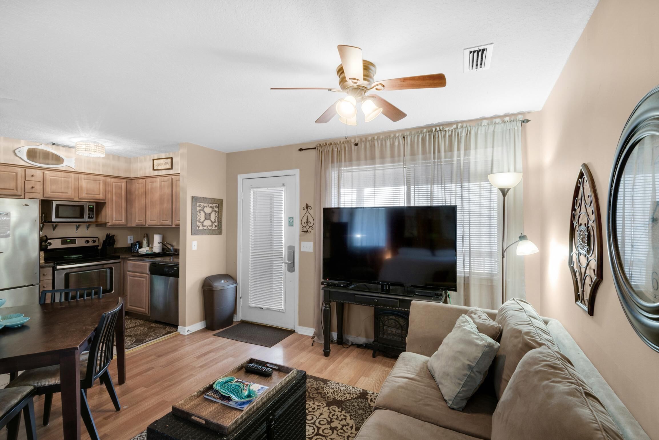 A modern living room with a flat-screen TV, a ceiling fan, and a dining area with wooden cabinets and a microwave.