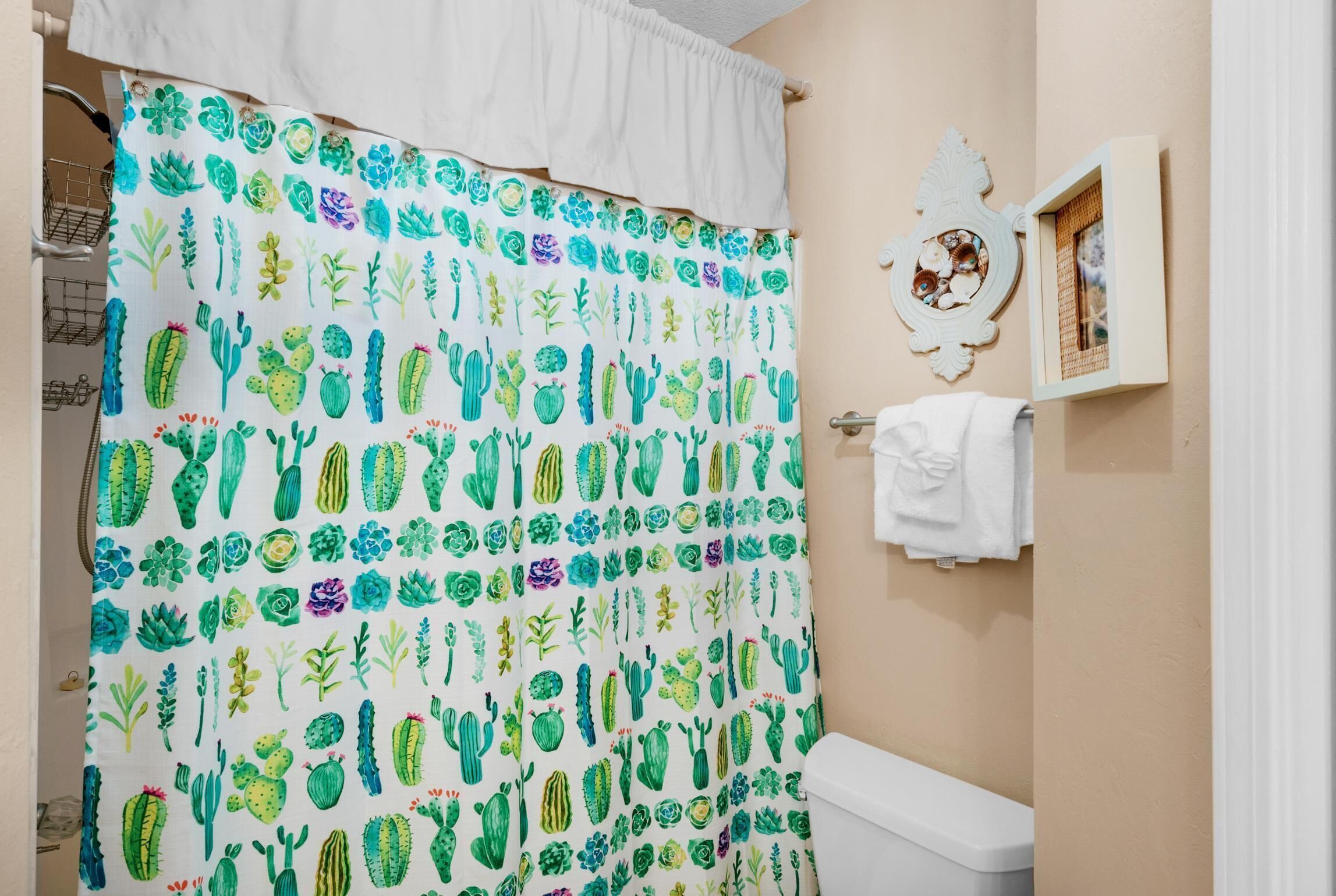Un baño con cortina de ducha estampada con motivos de cactus, un inodoro blanco y un cuadro en la pared.