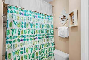 Un baño con cortina de ducha estampada con motivos de cactus, un inodoro blanco y un cuadro en la pared.