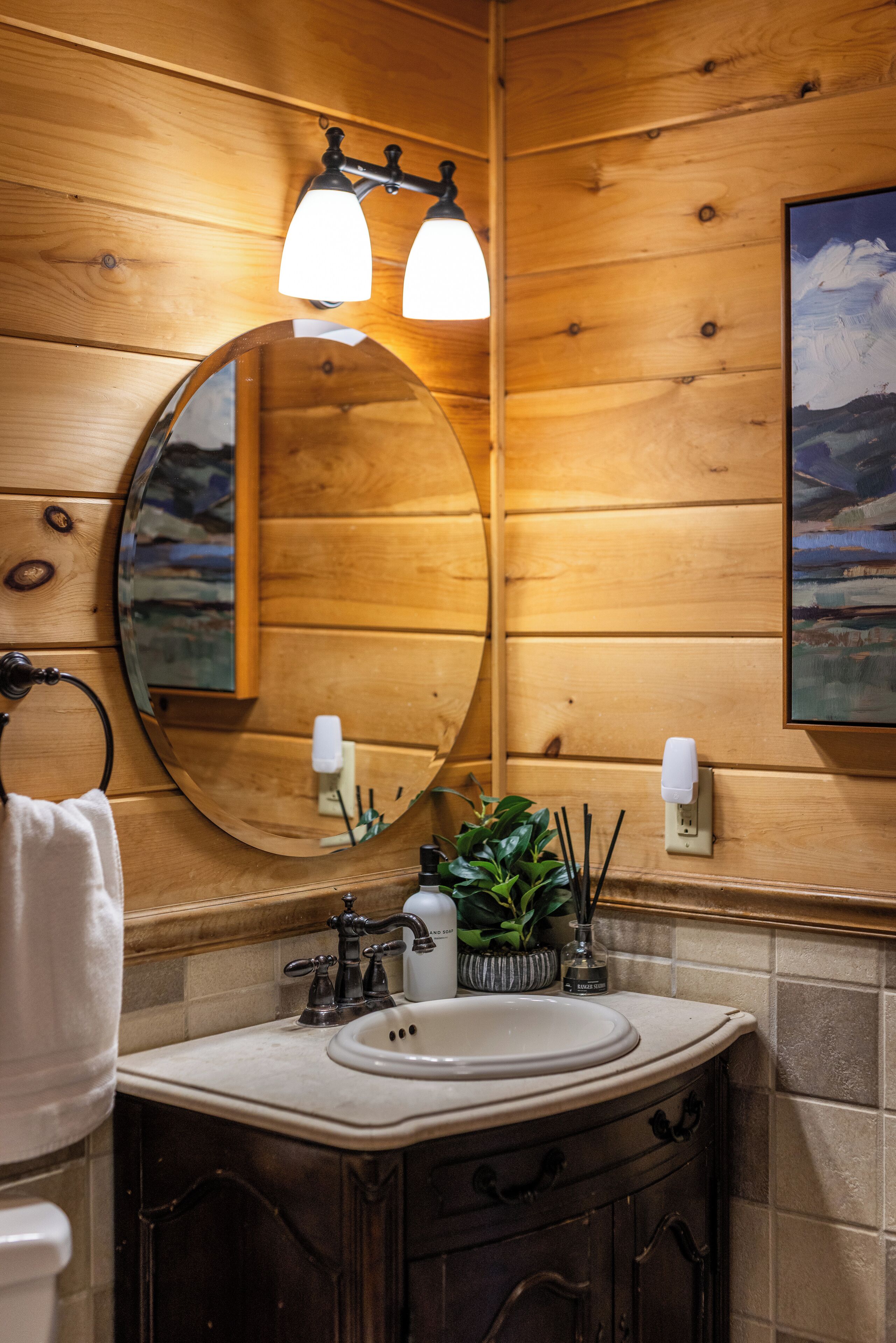 Une salle de bain de style rustique, avec des murs en bois, un miroir rond, deux appliques murales, un lavabo et une plante en pot.