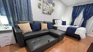 Une chambre à coucher moderne avec un canapé d’angle noir, un lit simple et une table de chevet avec une lampe.
