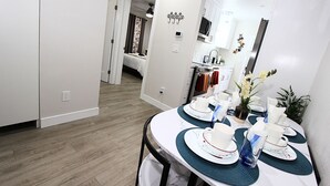 Un coin repas avec une table ronde dressée pour quatre personnes, une cuisine en arrière-plan et un couloir menant à une chambre.