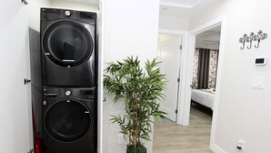 Une buanderie avec des lave-linge et des sèche-linge superposés, une plante en pot et une chambre à coucher visible à travers une porte ouverte.