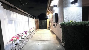 Un étroit sentier mène à une entrée fermée par un portail, de part et d’autre de laquelle s’étend une clôture blanche et une rangée de fleurs roses.