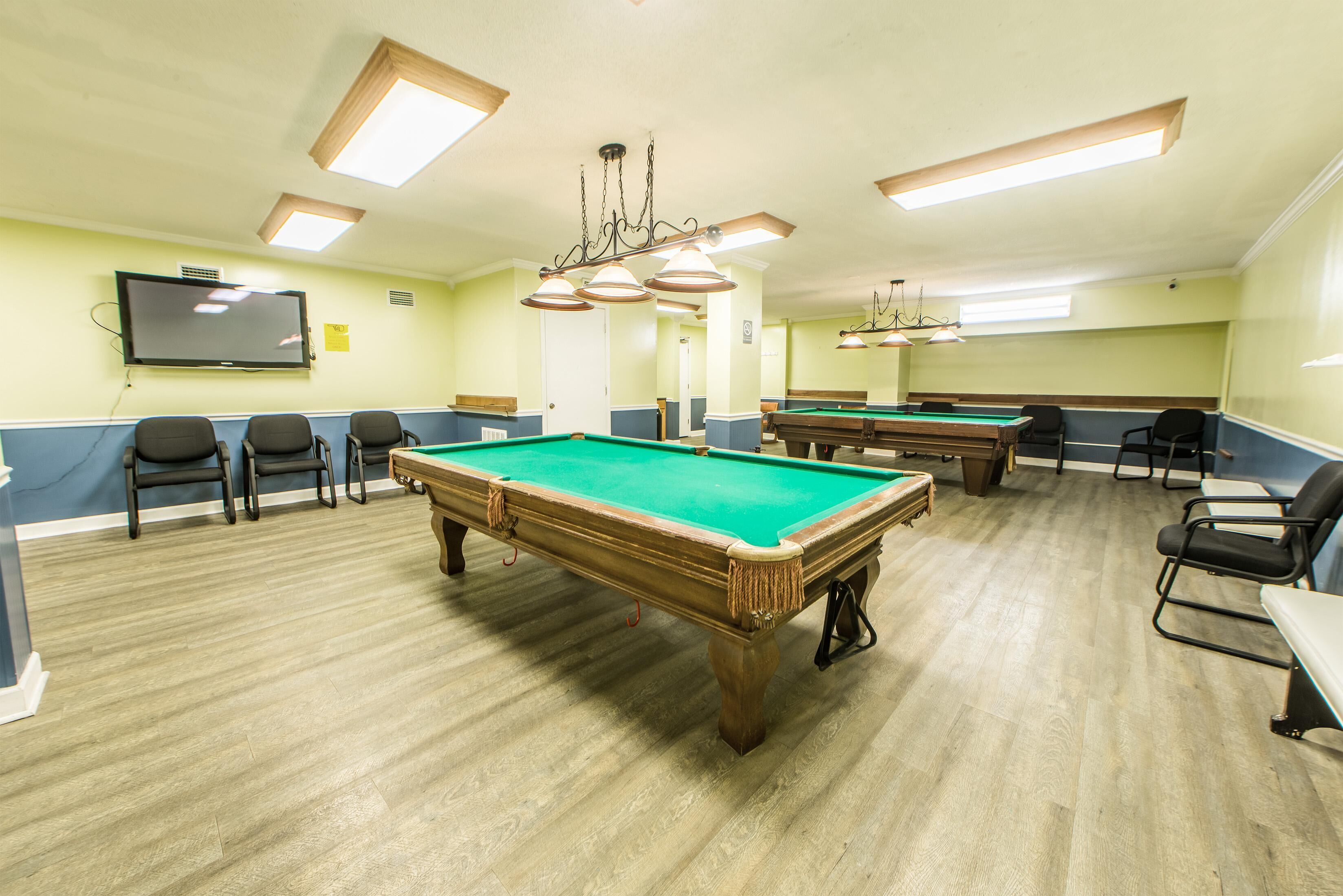 A pool room with multiple pool tables, chairs, and a television screen.