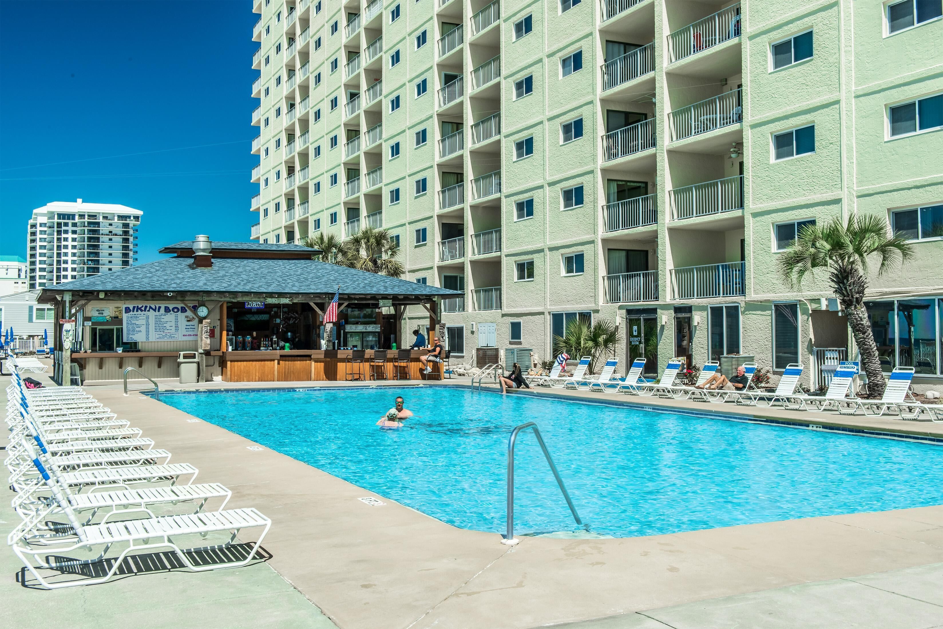 A hotel pool area with lounge chairs, a bar, and a building with multiple balconies.