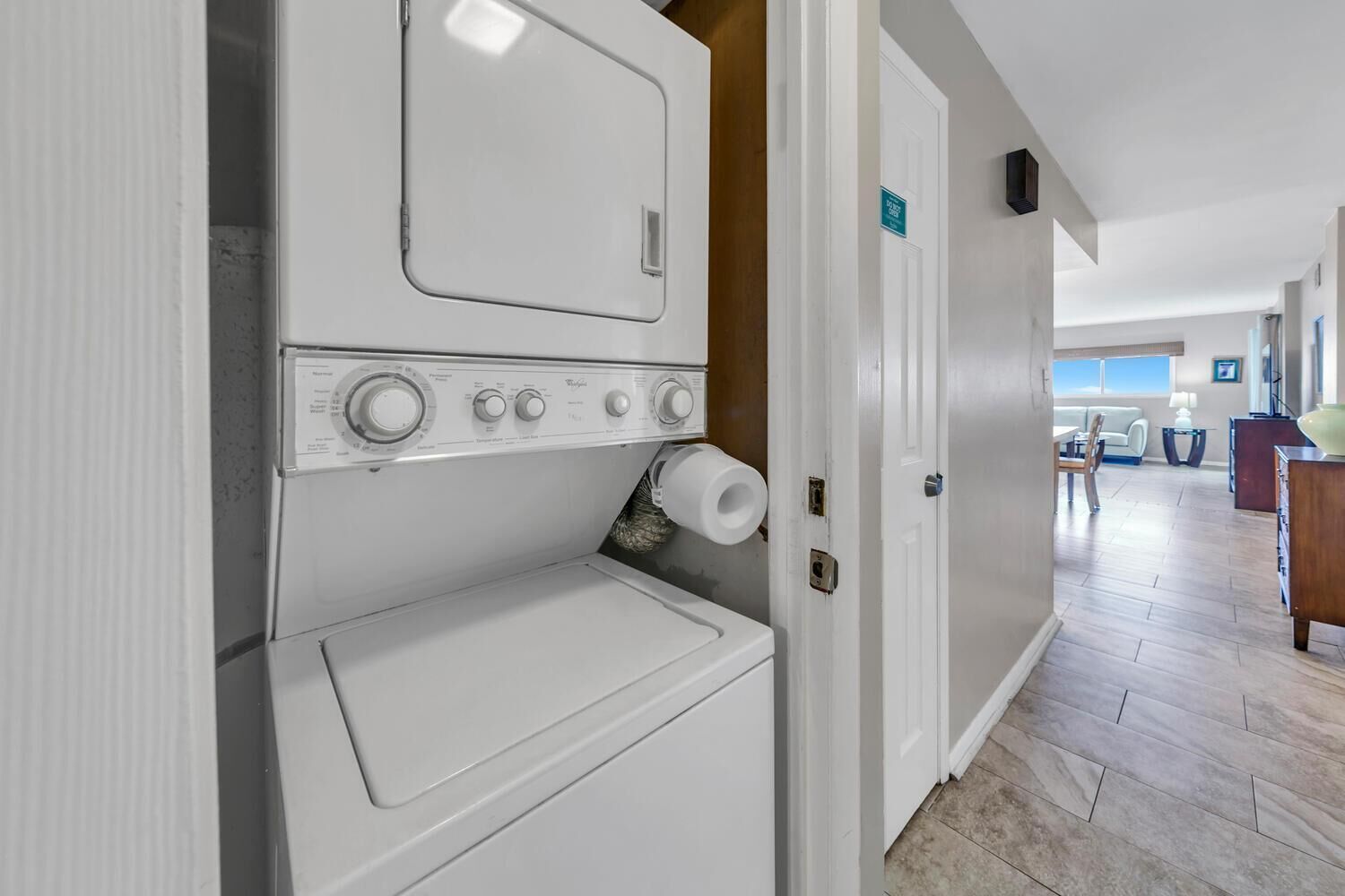 A laundry room with stacked washers and dryers, a door with a sign, and a view into a living area with a sofa and a table.