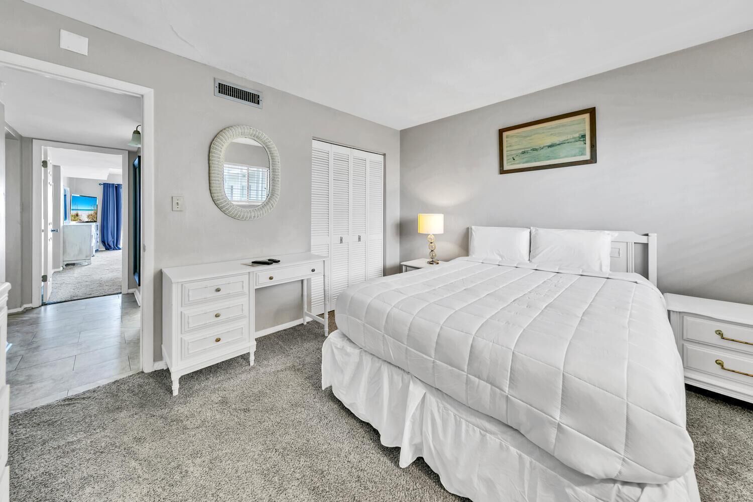 A bedroom with a bed, a dresser, a vanity with a mirror, a nightstand with a lamp, and a door leading to another room.