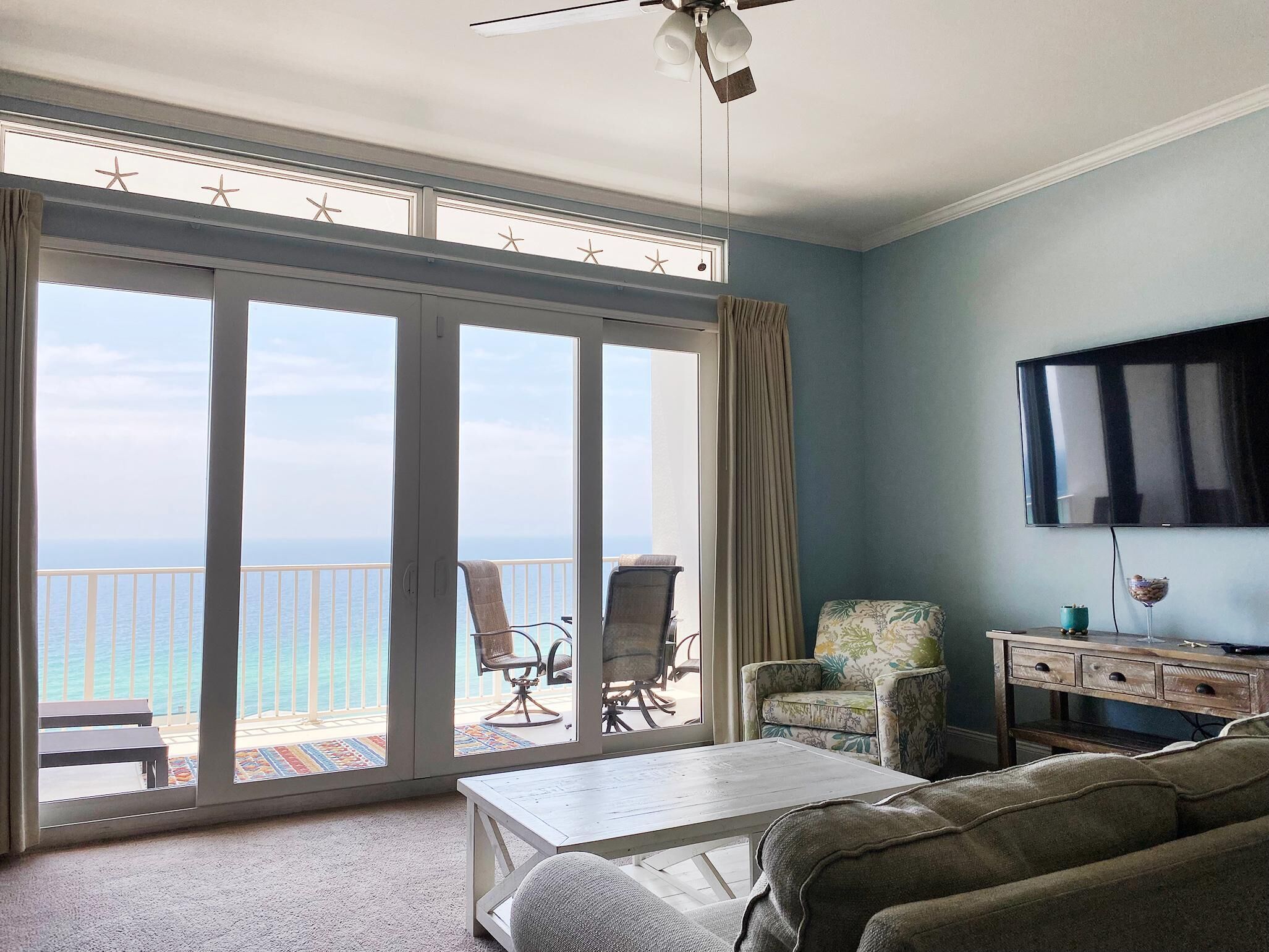 A living room with a view of the ocean, a ceiling fan, a TV, and a coffee table.