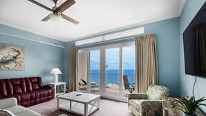 A spacious living room with a red leather sofa, a white coffee table, and a floral armchair. A ceiling fan is visible, and the room has a large sliding glass door leading to a balcony with a view of the ocean. - Laketown Wharf 2118 (Panama City Beach)