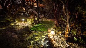 Un jardin nocturne doté d’une terrasse en bois, d’un chemin de pierre et de lanternes illuminées.
