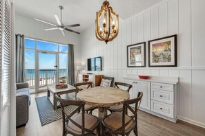 Un coin repas avec une table ronde en bois et des chaises, un ventilateur de plafond, un lustre et une vue sur la plage grâce à de grandes fenêtres.