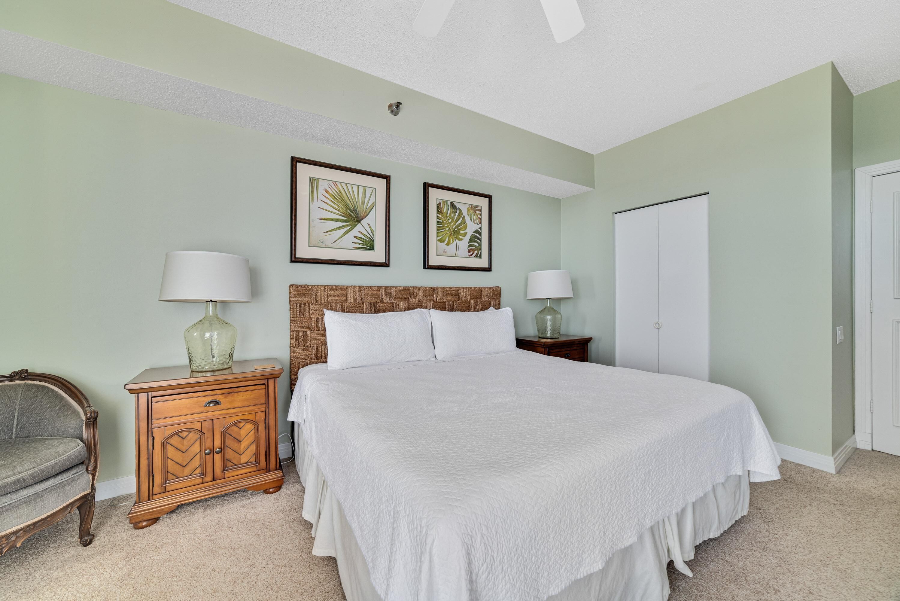 A bedroom with a large bed, two framed botanical prints on the wall, a nightstand with a lamp, and a wardrobe.