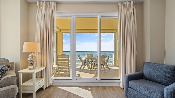 A room with a sofa, a table with a lamp, and a view of an outdoor patio with a table and chairs. - Beach Colony Tower 5B (Pensacola)