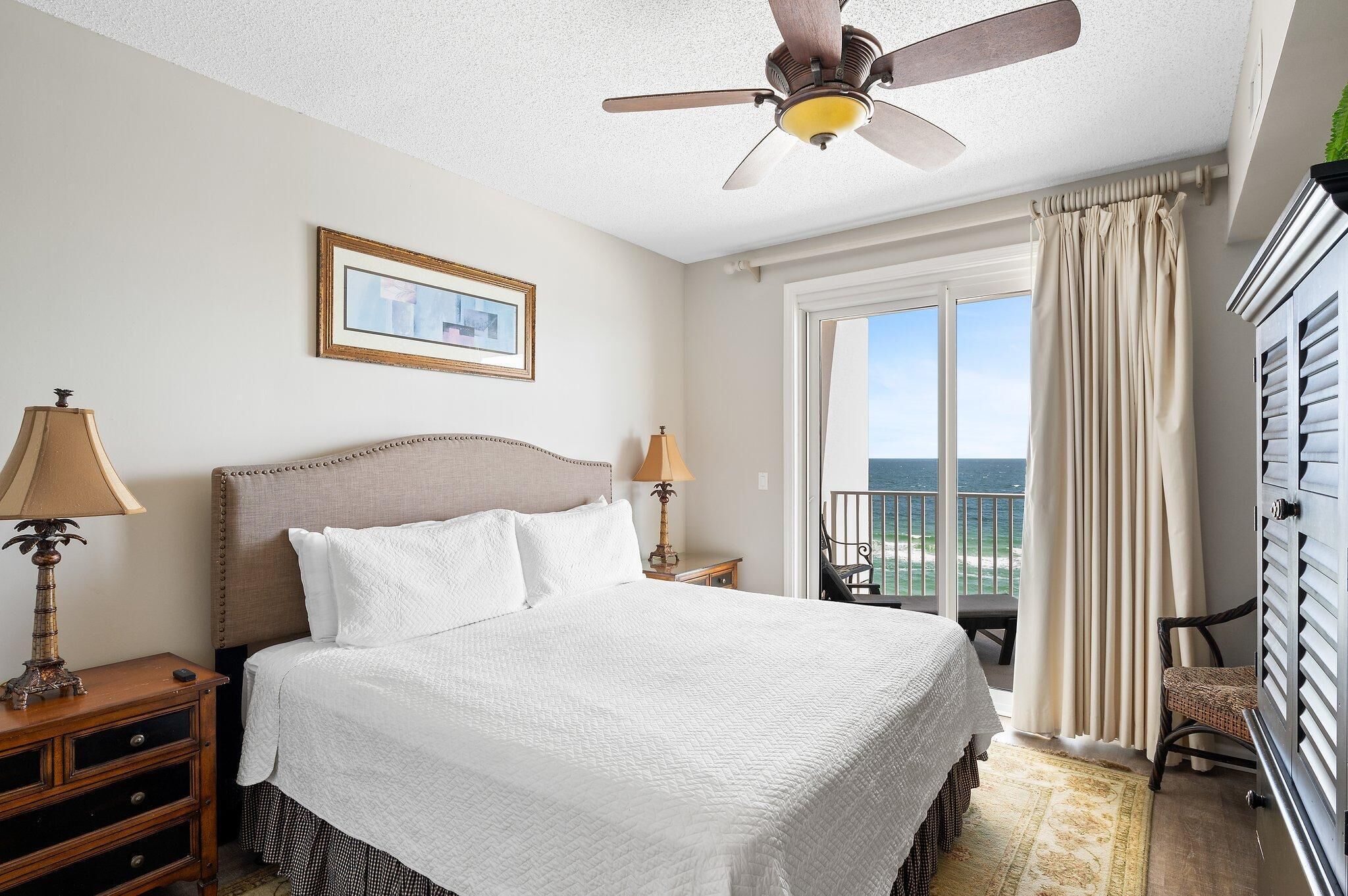 Ein Hotelzimmer mit einem großen Bett, zwei Nachttischlampen, einem Deckenventilator, einem gerahmten Bild an der Wand, einem Balkon mit Meerblick und einem Sessel am Fenster.