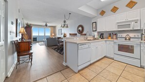 A modern kitchen with white cabinets, a central island, and a dining area with a chair and table. - Windemere 807 (Pensacola)